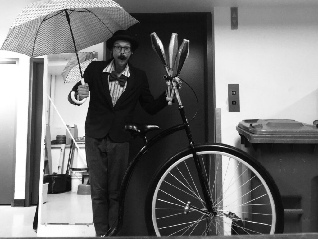 Homme avec un chapeau, moustache et habit, tenant un parapluie d'une main et des quilles et un velo à une roue de l'autre